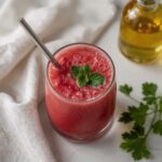 Glass of red how to detox your kidneys juice garnished with fresh herbs and served with a metal straw on a white surface.